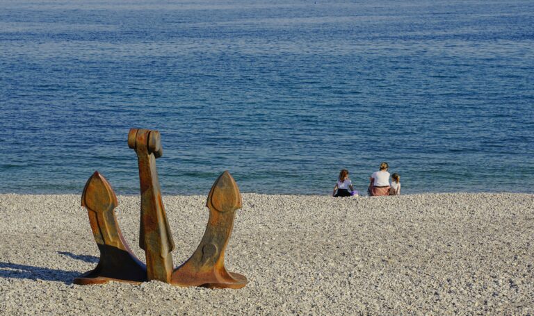 A rusty anchor on a pebble beach with a family enjoying the serene sea view.